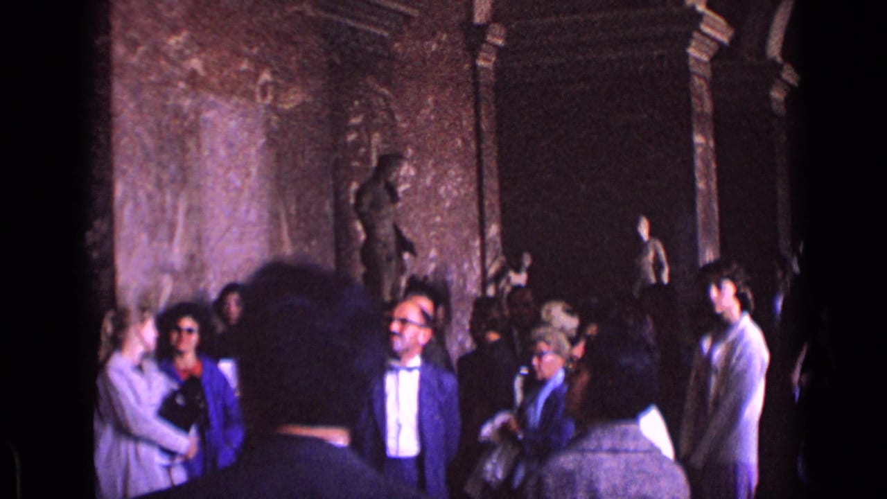 1962 Germany museum visitors stand beneath ornate walls near classical statues, with a suited man and bow tie near the center.