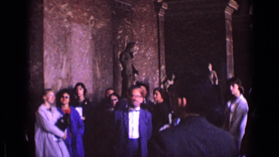 In a German museum gallery, adults in 1960s dress stand among columns and classical statues while another visitor is seen from behind.