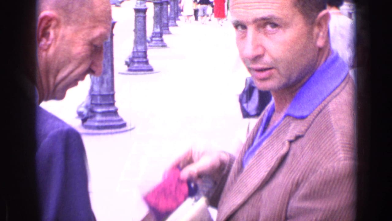 Two men stand close together on a paved street as one holds a small bright object.