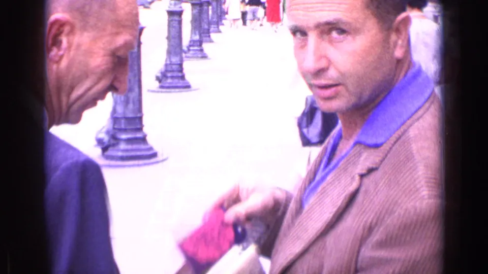 In 1962 archival home-movie footage, two adult men stand near cobblestones and lampposts while one handles a small pink and purple object.