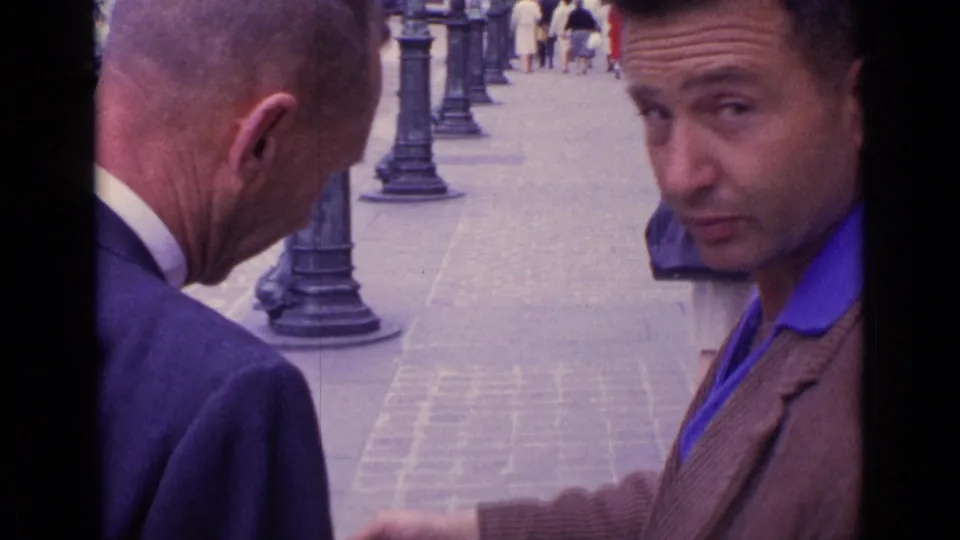 A close 1962 home-movie frame shows a man in a brown jacket glancing at the camera while an older man faces the street.