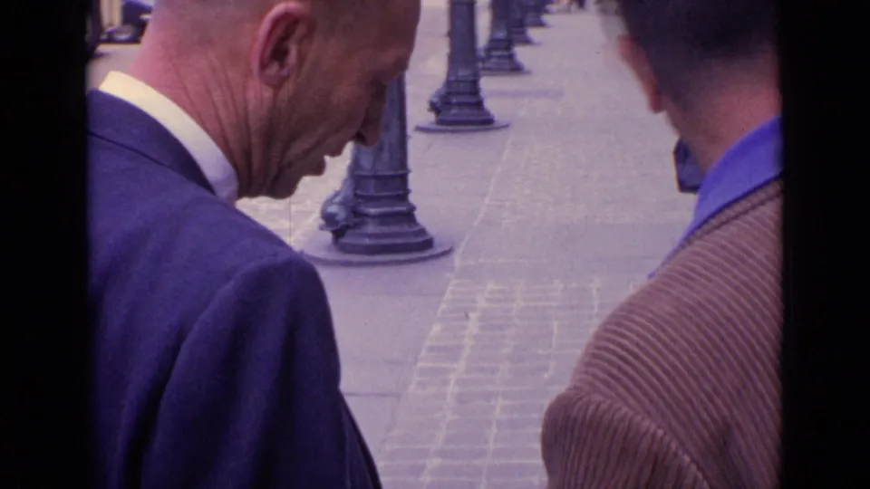 The home-movie camera frames the older man and the man in a brown jacket from close range on a paved European street.