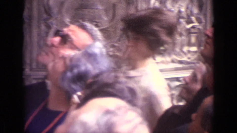 1960s tourists in Germany crowd near ornate cathedral carving, with one woman in glasses partly visible in profile.