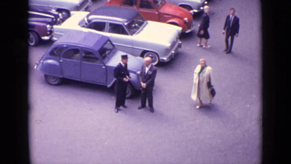 1962 Germany parking lot scene with classic European cars, a light blue small car, and adults in formal period clothing.