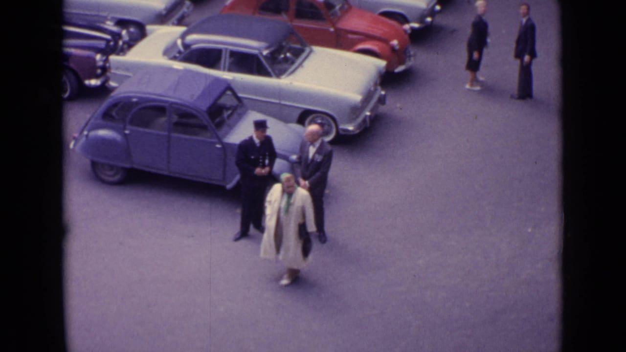 Woman in a light coat walks past men standing beside parked cars on a paved street.