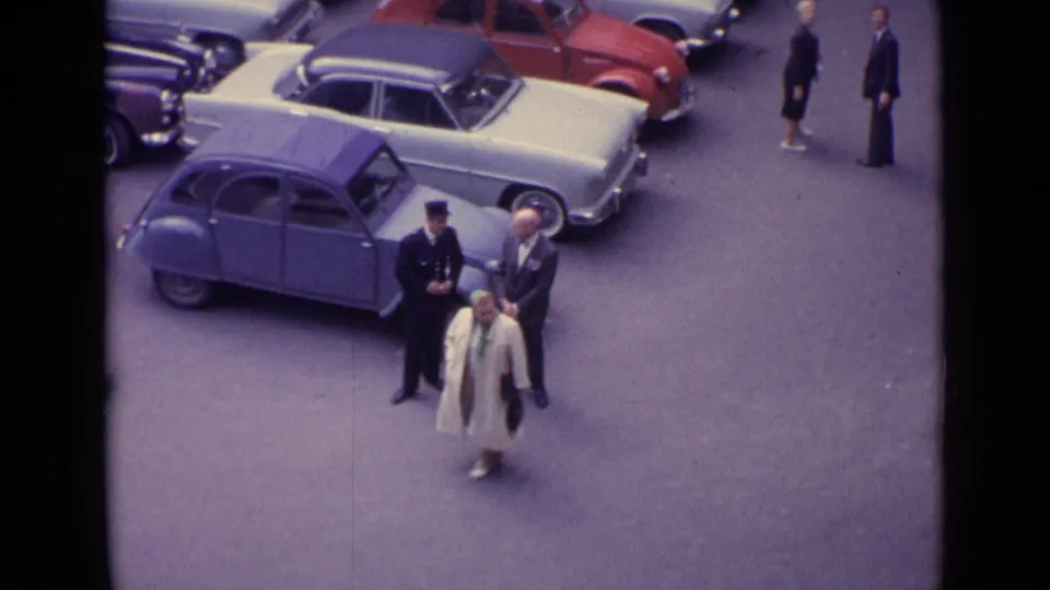 A woman in a light coat and headscarf passes two suited men and a uniformed man beside parked cars in a 1962 Germany street view.