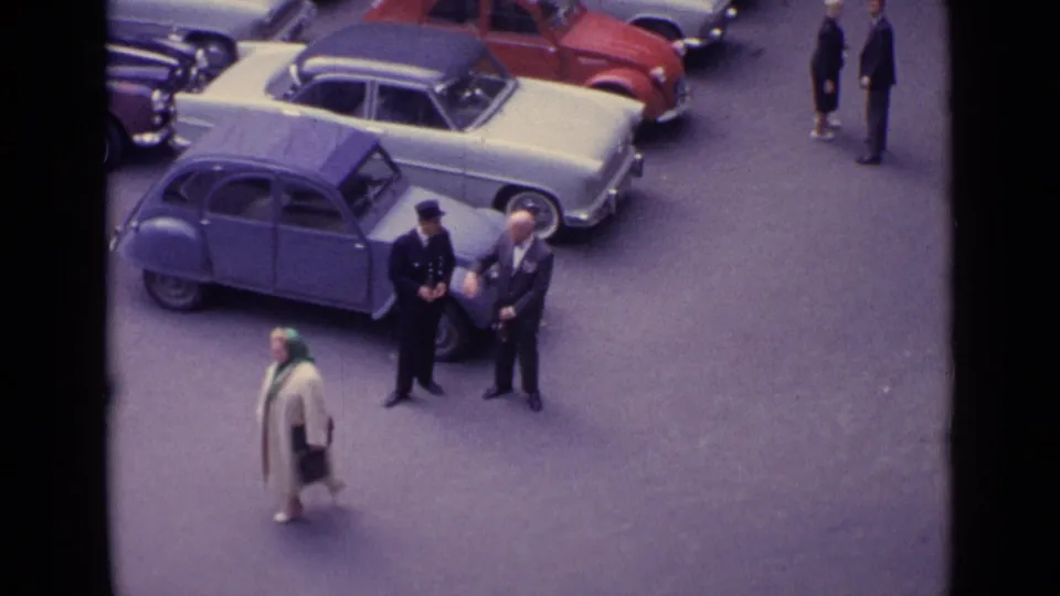 Elevated 8mm view of a woman moving through a German street scene as three men remain beside a blue compact car and other parked vehicles.