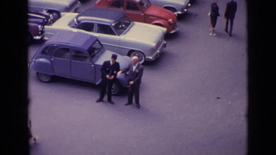 Three adult men remain by a blue compact car in a 1962 Germany street scene, with other pedestrians partly visible near the parked cars.