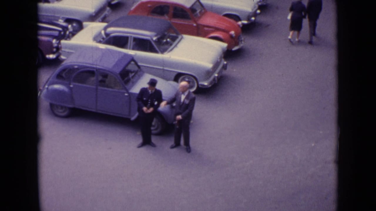 1962 Germany home-movie frame showing two men near a blue compact car in a busy parking area with mid-century automobiles.