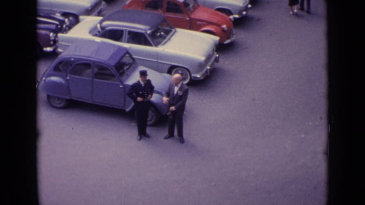 1962 Germany street-parking view with a uniformed man and suited man standing near a blue compact car among parked vehicles.