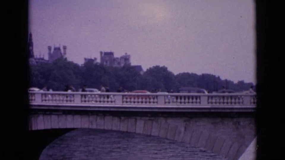 1962 Germany home-movie frame of a stone bridge over water, with pedestrians and period cars crossing under a hazy sky.