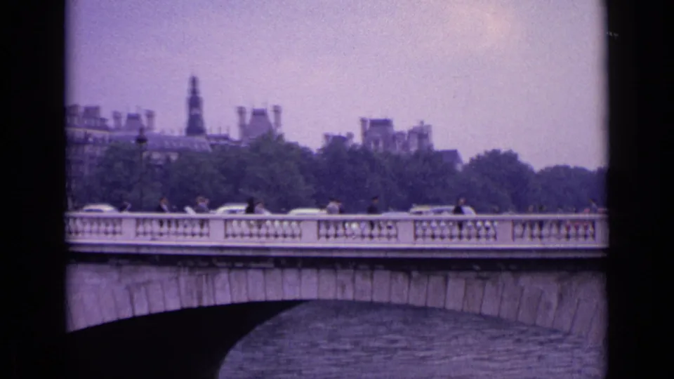 1962 Germany city view across a stone river bridge, with small figures, cars, trees, and distant rooflines visible.