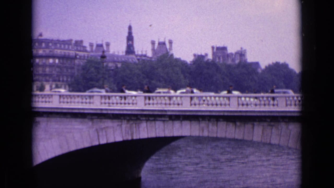 Stone arch bridge with cars and pedestrians crossing a river, with buildings and a spire beyond.