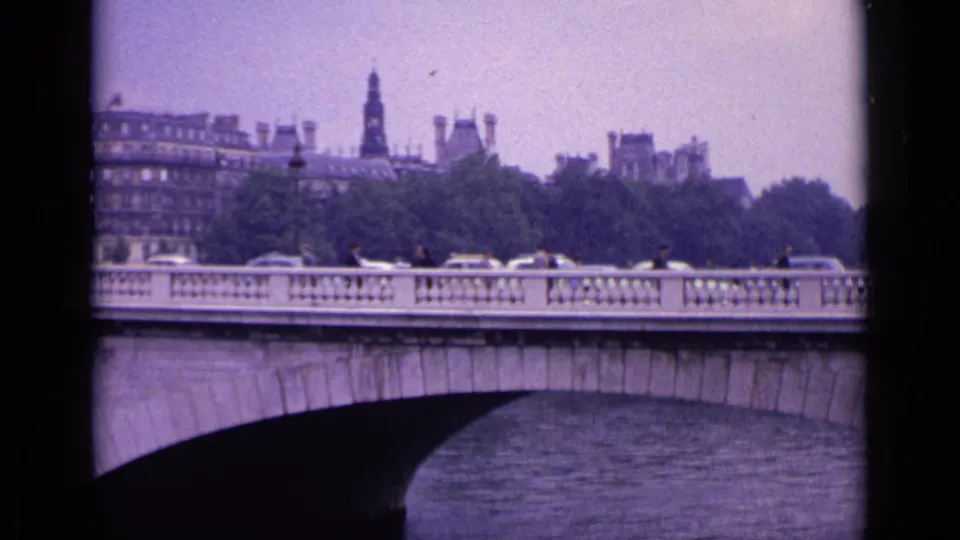 1962 8mm home-movie view identified as Germany, showing a stone bridge over water with traffic, pedestrians, trees, buildings, and a distant spire.