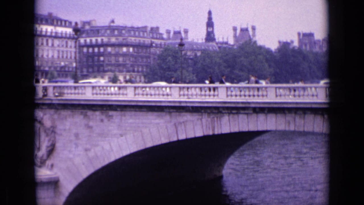 Bridge span over a river with cars and pedestrians, backed by trees, multi-story buildings, and a spire.