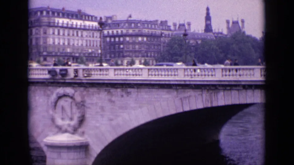1962 8mm home-movie frame identified as Germany, showing a stone bridge detail with an N medallion, passing traffic, pedestrians, and urban buildings.