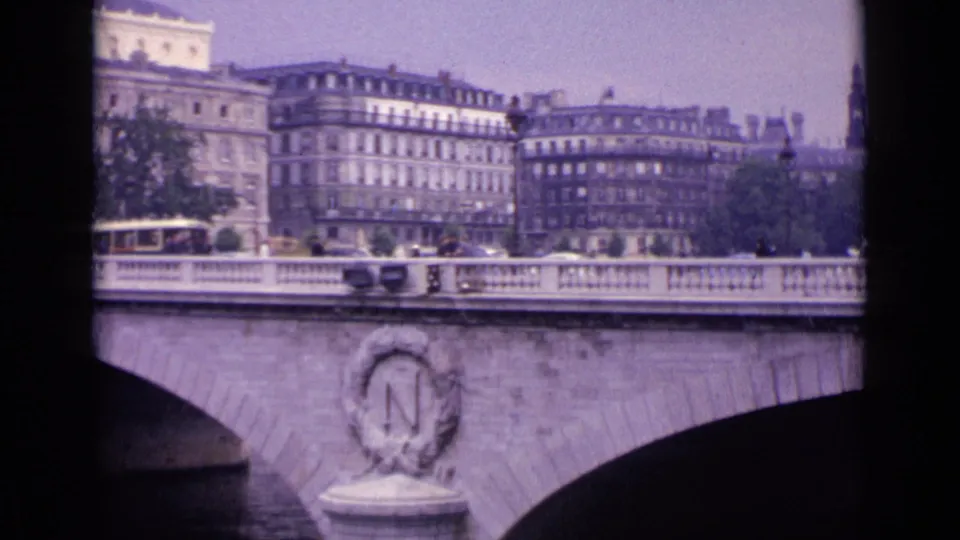 1962 8mm frame identified as Germany, showing the stone bridge facade, decorative N medallion, traffic, pedestrians, trees, and riverside buildings.