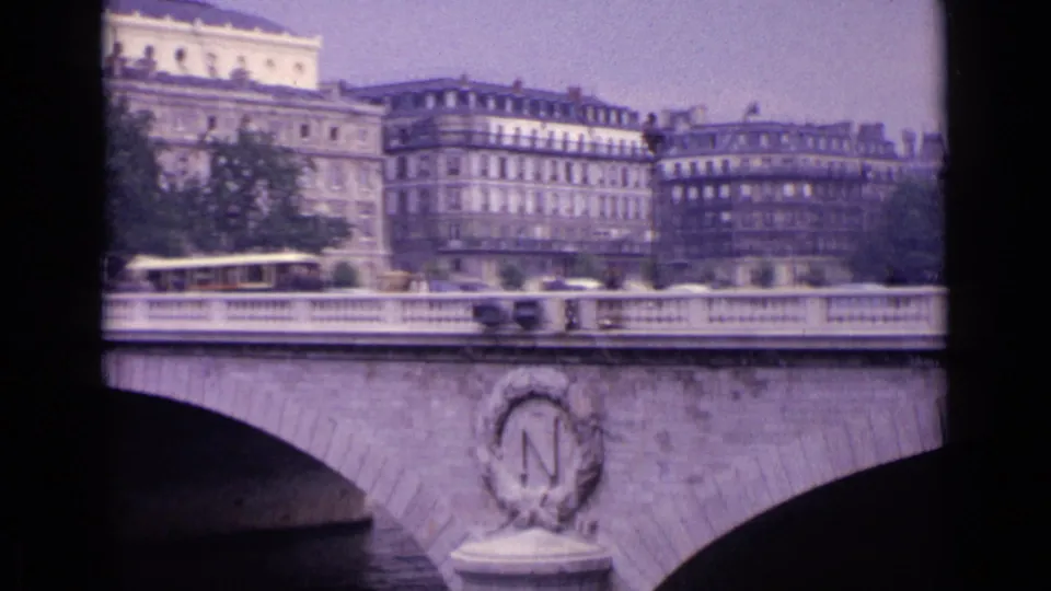 1962 8mm home-movie frame of a pale stone bridge, river arch, distant pedestrians, and multi-story European city buildings.