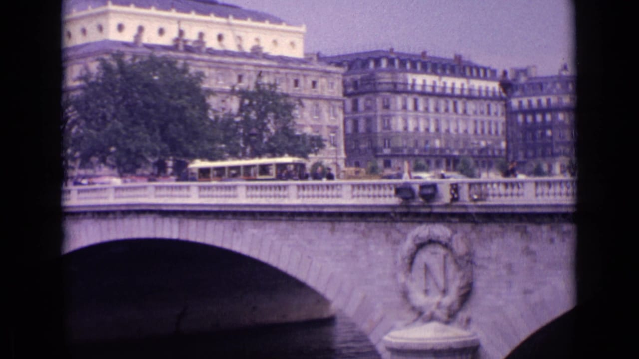 Soft 8mm view of a European river bridge with an oval stone emblem, street traffic, trees, and apartment-like buildings behind it.