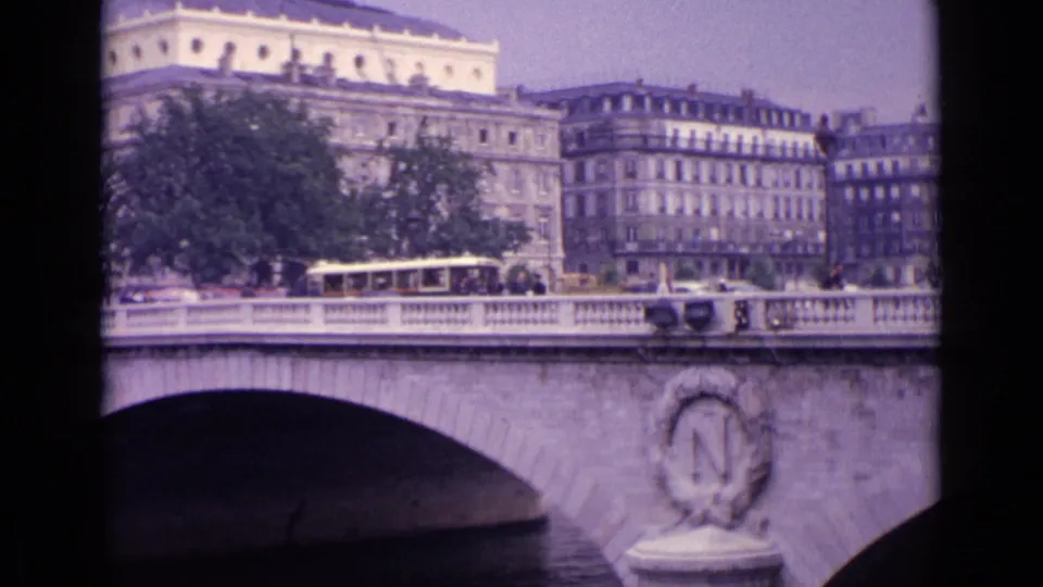 Soft 8mm view of a European river bridge with an oval stone emblem, street traffic, trees, and apartment-like buildings behind it.