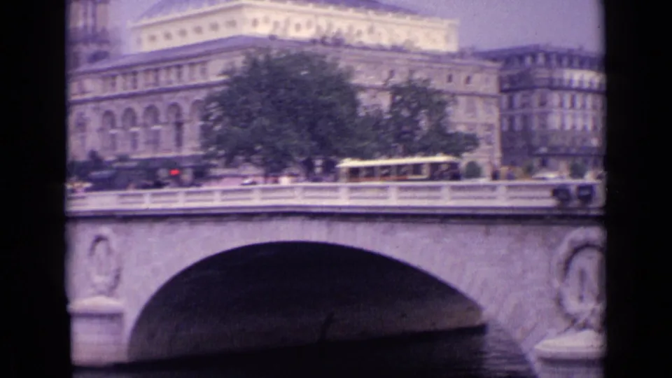 Vintage 8mm frame showing the bridge parapet, arched stonework, leafy trees, a yellow transit vehicle, and older urban buildings.