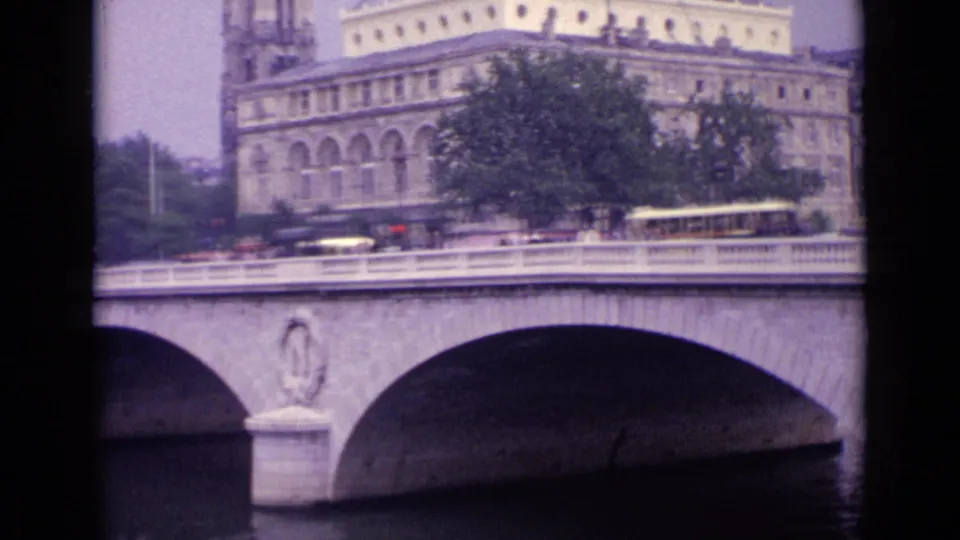 Final 8mm frame of a European stone bridge spanning dark river water, with traffic, pedestrians, trees, and urban buildings above.