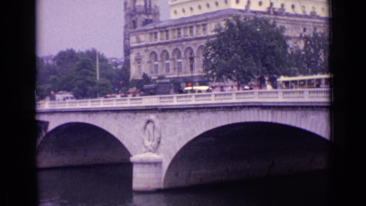 Stone arch bridge over a river with cars, a tram, trees, and a large classical building behind it.