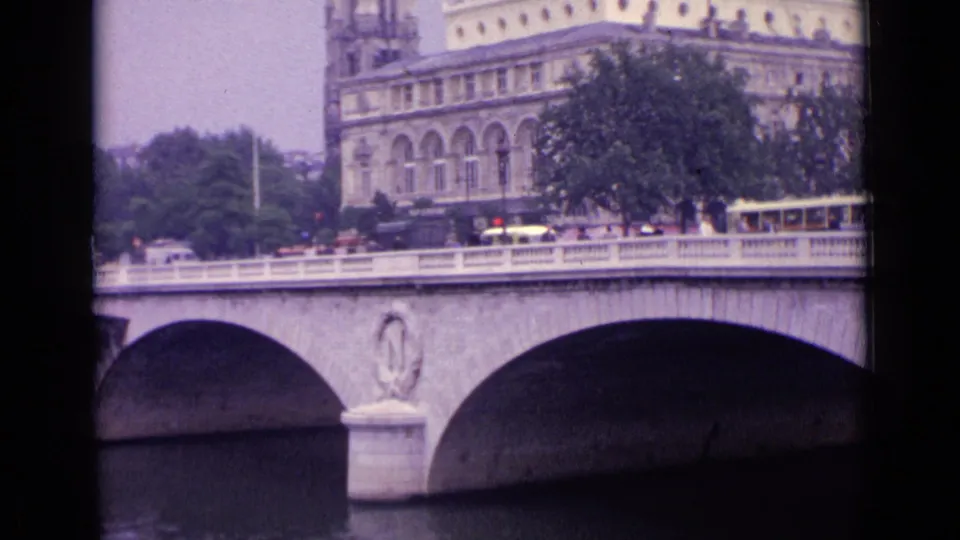 1962 8mm footage metadata places this public bridge scene in Germany, showing traffic, a tram, trees, and a grand building by the river.