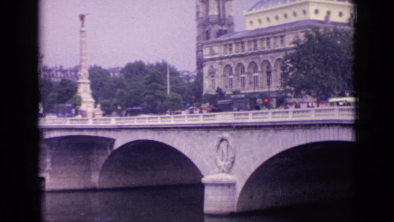 Bridge arches span the river with cars and a tram passing before trees and a large building.