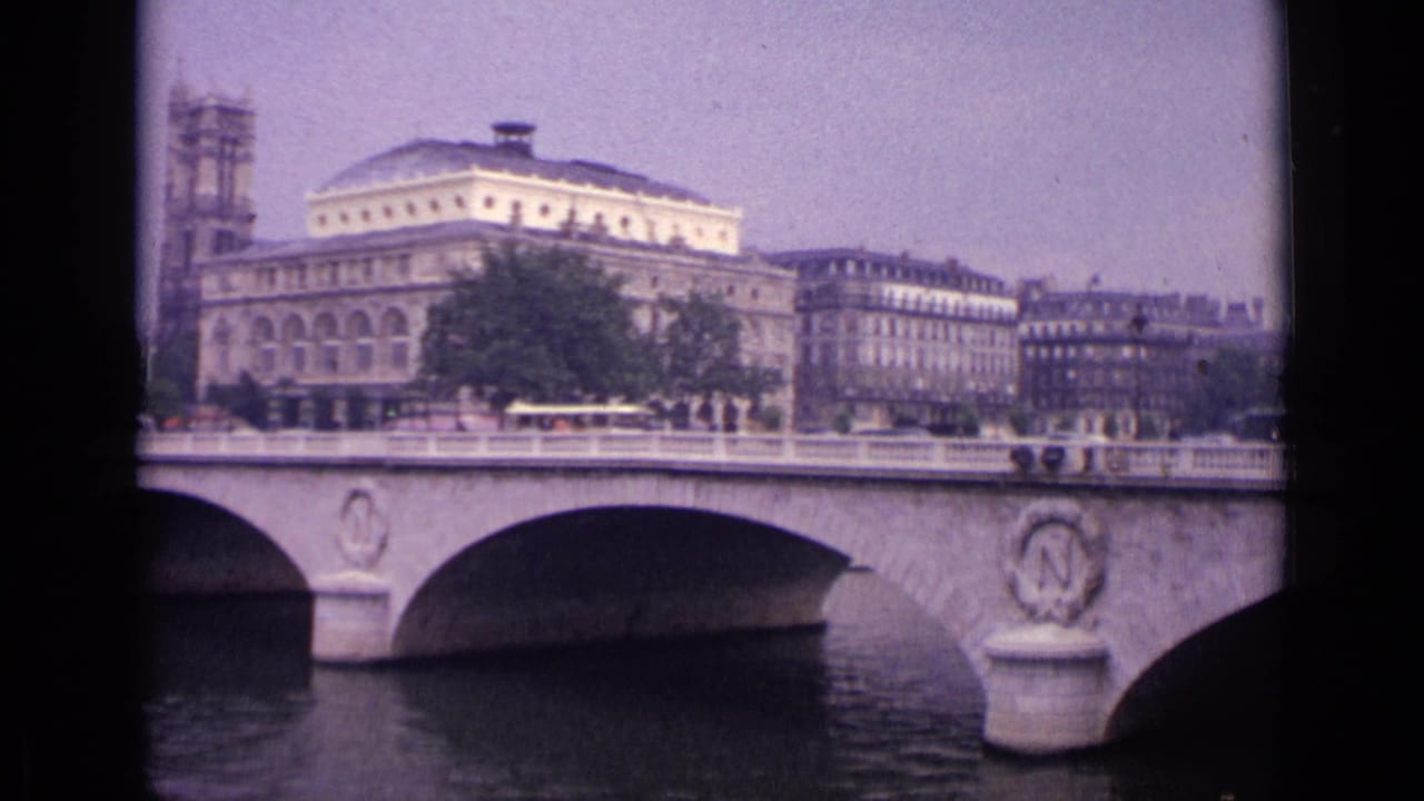 1962 8mm view in Germany of a stone bridge over a river, with pedestrians, vehicles, trees, and formal urban buildings beyond.