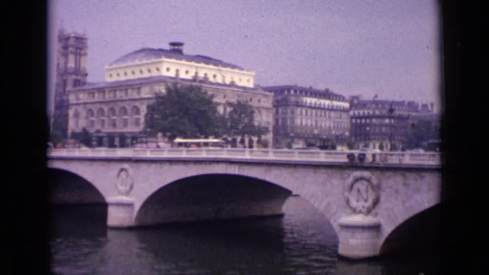 1962 8mm view in Germany of a stone bridge over a river, with pedestrians, vehicles, trees, and formal urban buildings beyond.