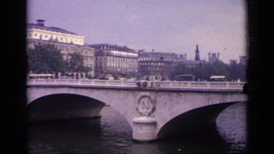 1962 German city bridge scene with cars or buses crossing, a stone arch over the river, and multi-story buildings in the background.