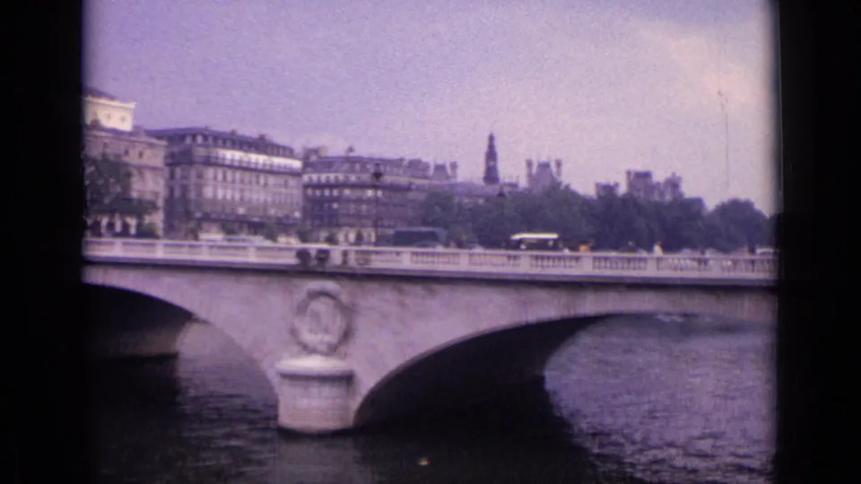 The 8mm frame shows a stone bridge with an ornamental medallion, small vehicles, trees, and distant buildings in a German city setting.