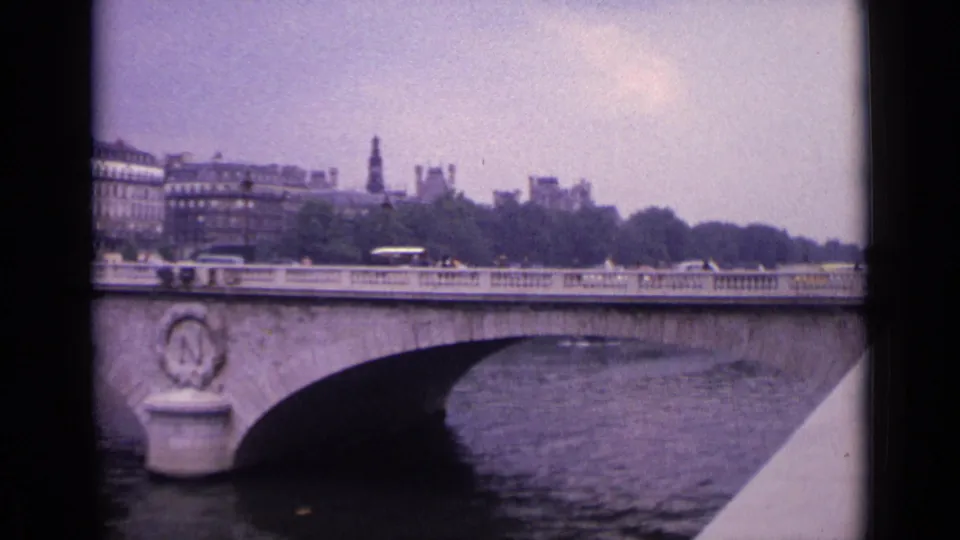 1962 8mm view across a German river bridge, showing traffic on the deck, a broad arch, trees, and hazy city buildings in the distance.