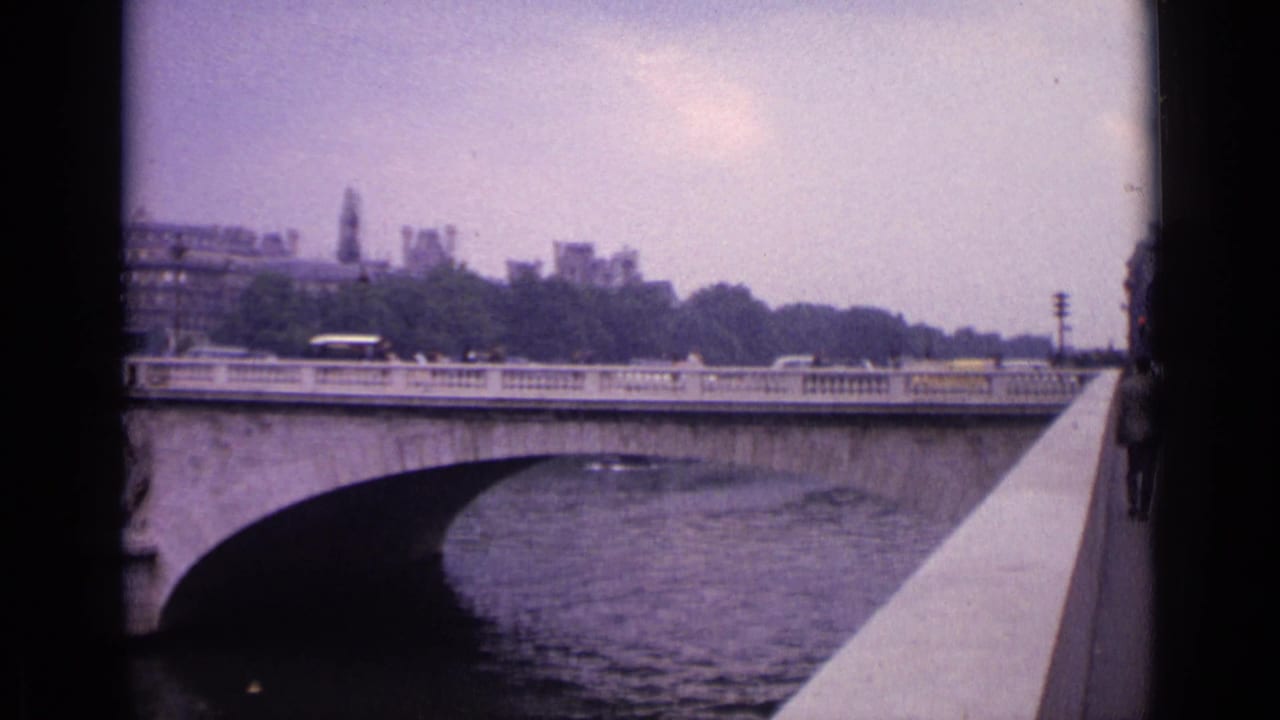 1962 Germany 8mm frame showing a stone bridge over a river, traffic on the bridge, and a riverside parapet from a handheld viewpoint.