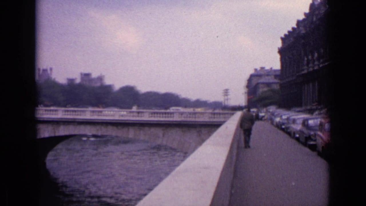 1962 Germany 8mm frame looking down a riverside sidewalk with parked classic cars, a pedestrian, river wall, and masonry buildings.