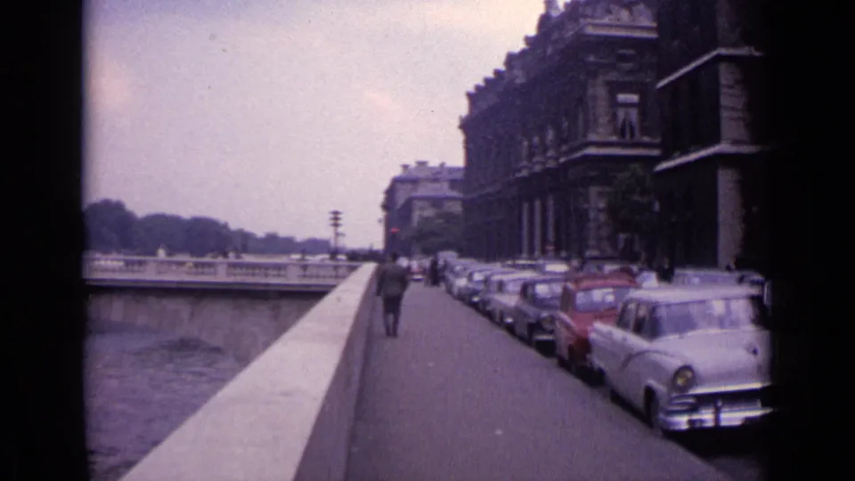 1962 Germany street view with parked classic cars, a pedestrian walking ahead, a river parapet, and large urban buildings.