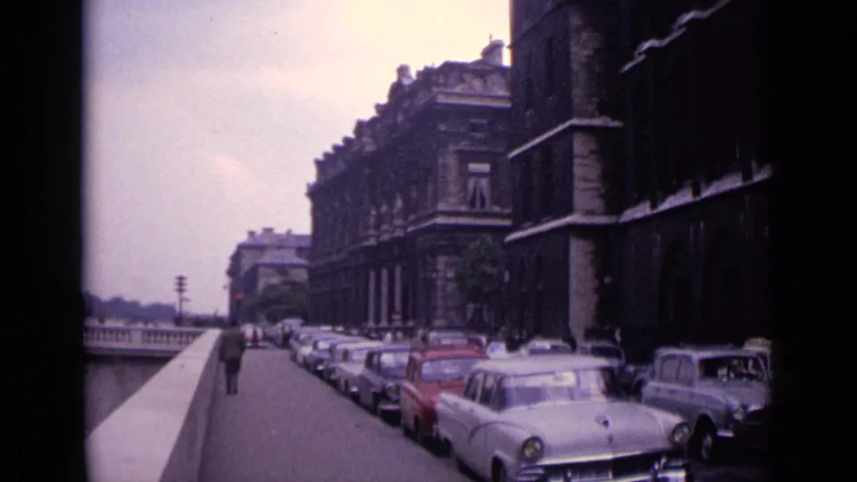 1962 Germany 8mm frame of a riverside street with rows of parked cars, pedestrians in the distance, and formal masonry buildings.