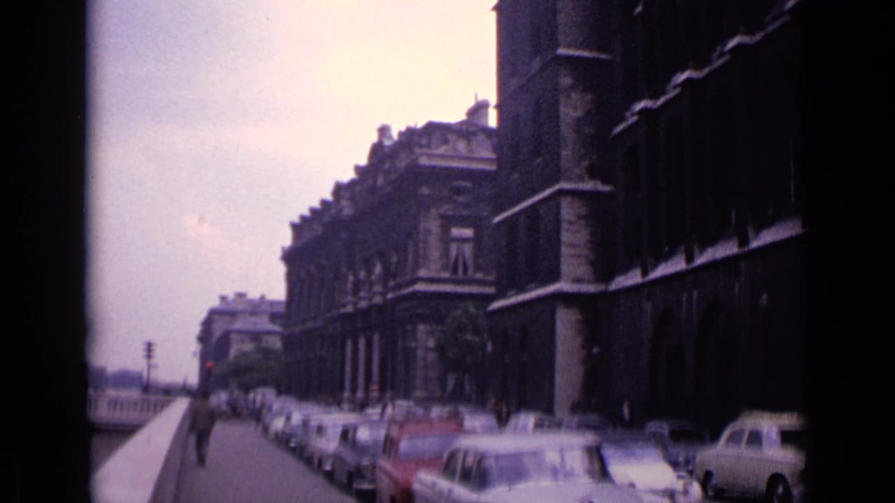 1962 Germany 8mm frame showing parked cars along a stone-edged street beside historic urban facades.
