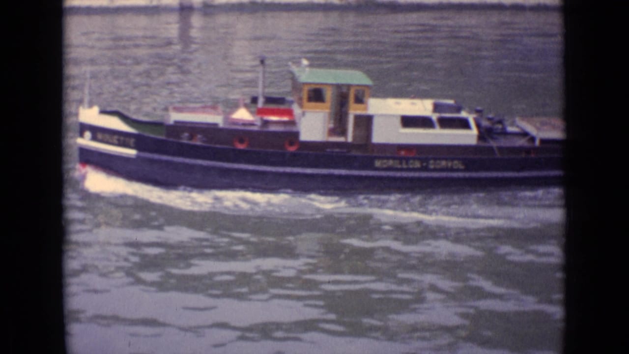 A small marked tugboat travels across a German river in faded 1962 8mm home-movie footage, with cabin, hull lettering, and wake visible.