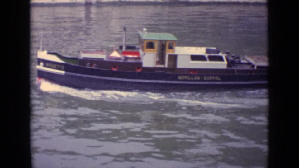 A small marked tugboat travels across a German river in faded 1962 8mm home-movie footage, with cabin, hull lettering, and wake visible.