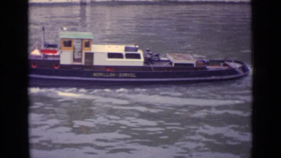 Side view of the small tugboat on the river, with its cabin, low deck, painted hull markings, and rippled water visible in the 8mm frame.