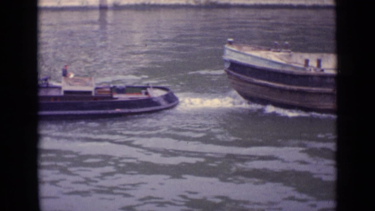 1962 Germany 8mm frame showing a small tugboat pulling near the bow of an empty barge on a river.