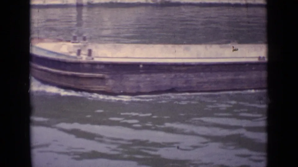 1962 Germany 8mm frame showing the side of a large empty barge moving past on rippling water.