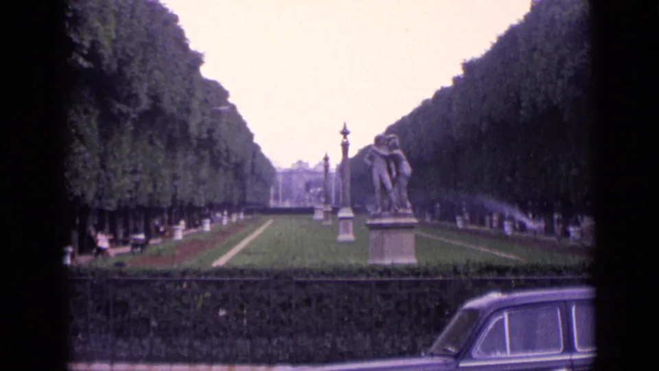1962 German park scene showing a formal lawn, clipped tree avenues, sprinkler spray, statue, benches, fence, and the rear of a vintage car.