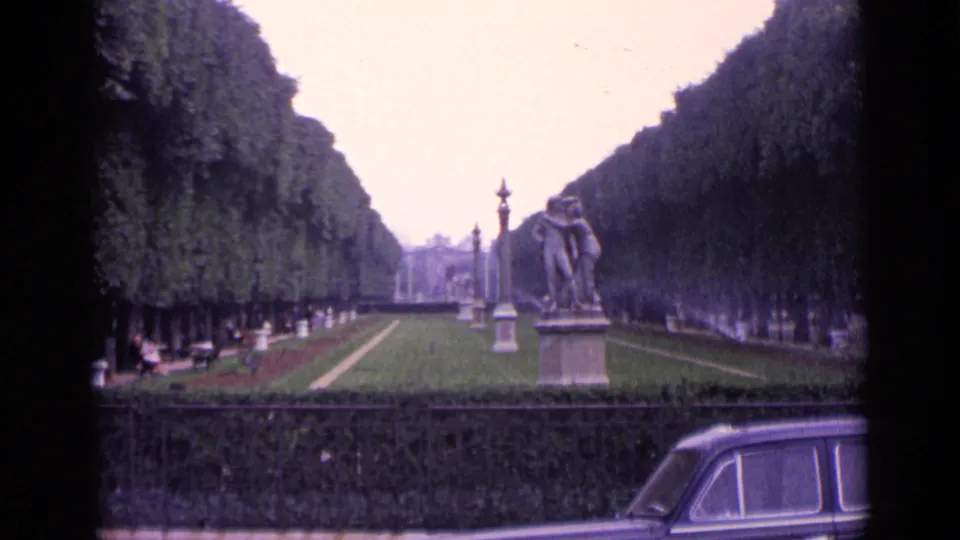 1962 Germany home-movie frame of a symmetrical park avenue with clipped trees, lawn sprinklers, a statue, fence, benches, and a partial car.
