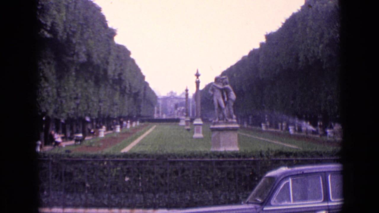 1962 Germany home-movie frame showing a formal park avenue with statues, clipped trees, sprinkler spray, and a classic car crossing the foreground.