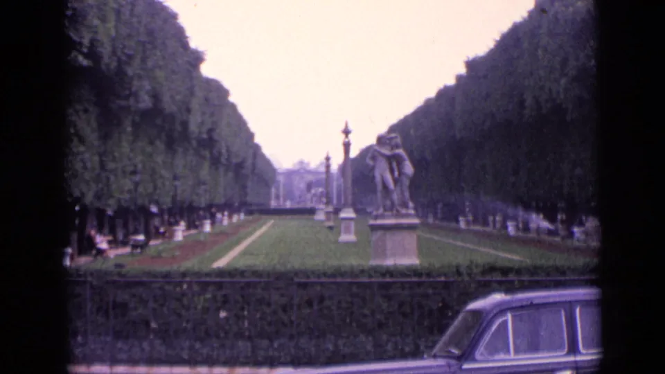1962 Germany home-movie frame showing a formal park avenue with statues, clipped trees, sprinkler spray, and a classic car crossing the foreground.