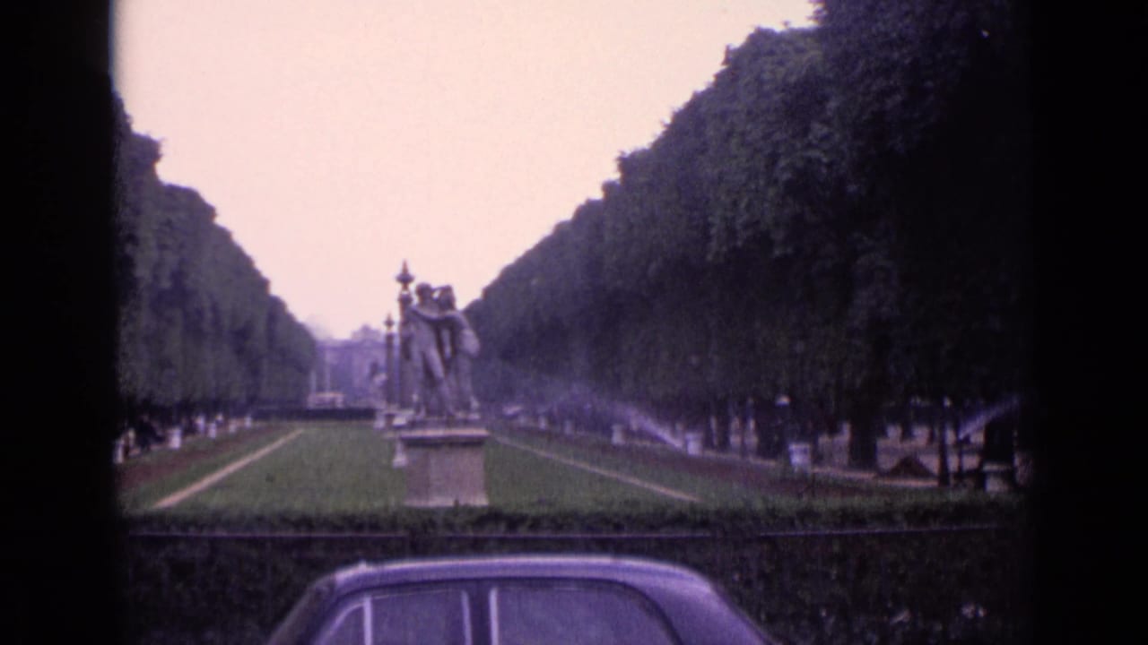 1962 Germany park scene with a long formal avenue, pedestal statues, sprinkler mist, distant visitors, and the roofline of a classic car in the foreground.