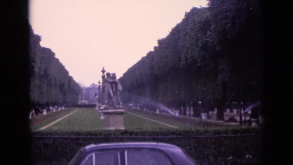 1962 Germany park scene with a long formal avenue, pedestal statues, sprinkler mist, distant visitors, and the roofline of a classic car in the foreground.
